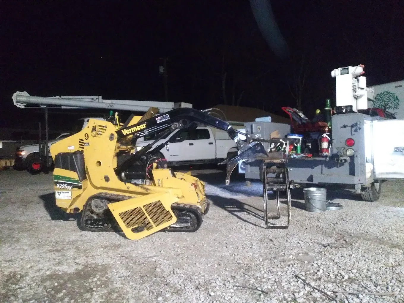 Yellow skid steer loader parked next to a work truck under nighttime lighting.
